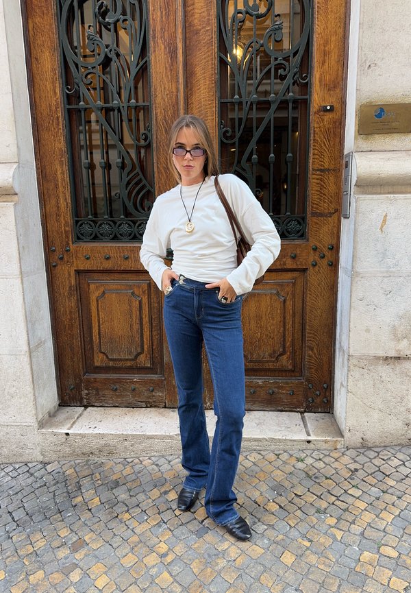 Mujer con camiseta blanca de manga larga, vaqueros azules, zapatos negros y gafas moradas, de pie con las manos en los bolsillos delante de puertas dobles de madera.