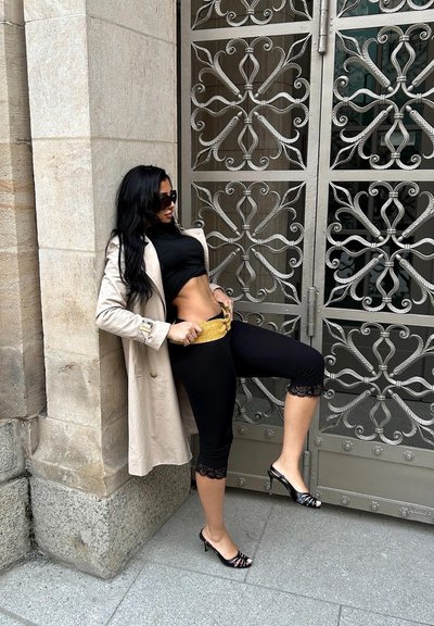 Mujer con largo cabello negro, con gafas de sol, abrigo beige, top corto negro y mallas, ajustando un cinturón dorado junto a una puerta de metal ornamentada.