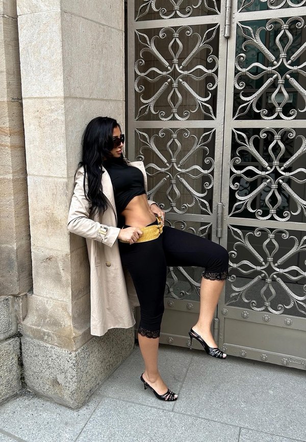 Frau mit langen schwarzen Haaren, die eine Sonnenbrille, einen beigen Mantel, ein schwarzes kurzes Oberteil und Leggings trägt, richtet einen goldenen Gürtel an einem verzierten Metalltor.