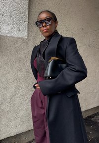 Femme confiante portant de grandes lunettes de soleil noires, un manteau noir, une tenue bordeaux, tenant un sac à main noir, debout contre un mur beige texturé.