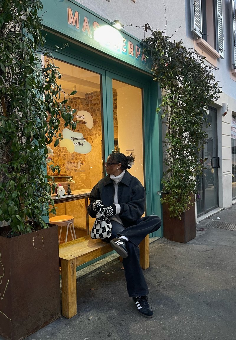 Extérieur d'un café spécialisé avec un cadre en teal, un banc en bois et des plantes en pot. Une personne en tenue noire est assise avec un sac à carreaux et des gants.