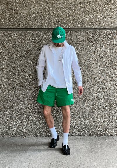 Camisa blanca de botones sobre una camiseta blanca, pantalones cortos verdes, gorra verde, zapatos negros y calcetines blancos, de pie contra una pared gris texturizada.