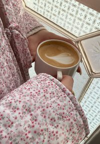 Hvid kaffekop med latteart holdt i en hånd, iført et let blomsterprintet tøj med pink og grønne accenter.