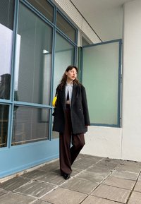 Μαύρο παλτό από μαλλί με υφή, γκρι ριγέ μπλούζα και καφέ φαρδύ παντελόνι. Κίτρινη τσάντα ως ένδειξη αντίθεσης; σύγχρονος, μινιμαλιστικός σχεδιασμός.