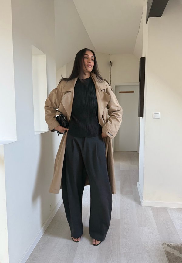 Beige trench coat over a fitted black ribbed jumper, paired with loose black trousers and black open-toed sandals. Simple, modern design.