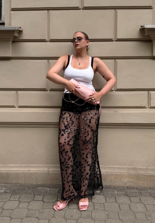 Frau trägt weißes Tanktop, schwarzen durchsichtigen floralen Rock über Shorts, pinke Sandalen und hält eine pinke Clutch, steht vor einer beigen Wand.