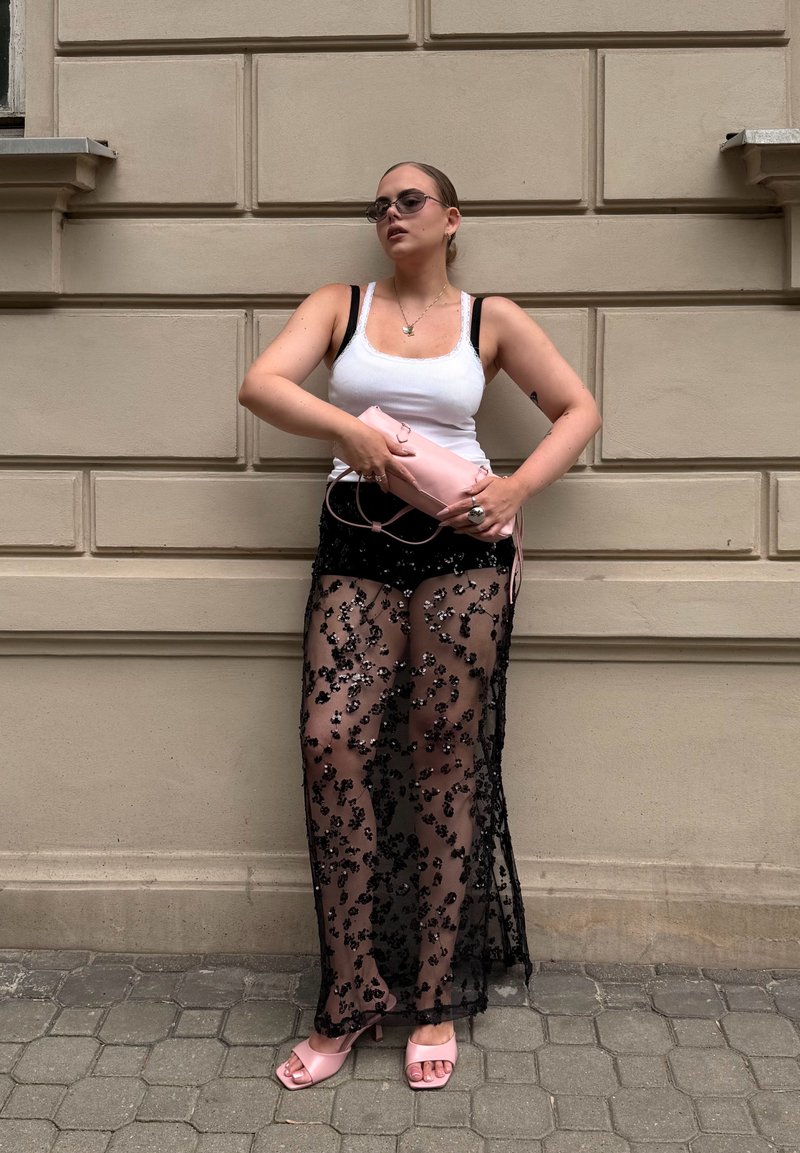 Femme portant un débardeur blanc, jupe fleurie noire transparente par-dessus un short, sandales roses tenant une pochette rose, debout contre un mur beige.