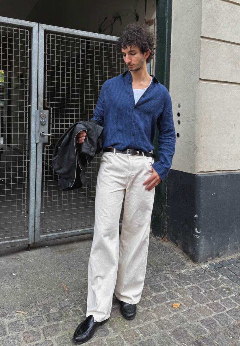 Camisa azul riscada com botões, calças brancas de perna larga, jaqueta de couro preta pendurada no braço e loafers pretos. Cenário urbano com portões de metal.