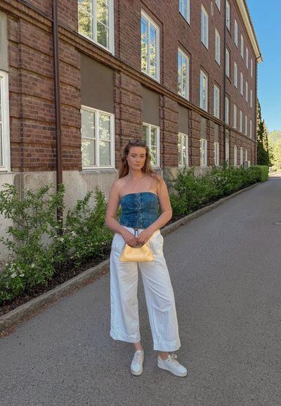 Joven mujer está de pie fuera de un edificio de ladrillo, vestida con un top azul sin tirantes, pantalones blancos, zapatillas blancas y sosteniendo un pequeño bolso amarillo.