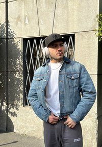 Chaqueta de mezclilla con un lavado de azul claro, llevada sobre una camiseta blanca. Pantalones de chándal gris oscuro con un número impreso, auriculares blancos y una gorra negra.