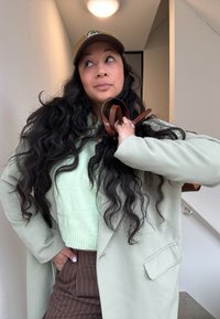 Groene fleecjas over een lichtgroene trui, gecombineerd met bruine gepinstripete broeken. Houdt bruine leren banden vast, met lang golvend haar.