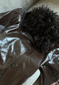 Hnedá kožená bunda s kučeravým čiernym kožušinovým golierom, veľkými gombíkmi a hladkým, lesklým povrchom. Strihaný strih so stranovými vreckami.