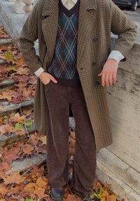 Brown checked long coat over a dark sweater with an argyle pattern, paired with brown corduroy trousers and black shoes, on a leaf-covered ground.