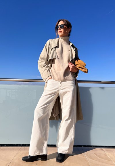 Suéter de punto beige, abrigo de trench gris claro, pantalones anchos blancos, botas de tacón negras y accesorio marrón; todo contra un cielo azul claro.