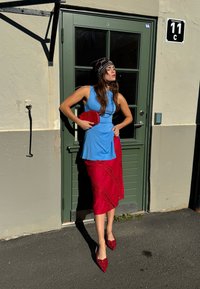 Top sem mangas azul claro, saia vermelha com detalhes intrincados em renda, sapatos de verniz vermelho e uma pequena bolsa vermelha; de pé ao lado de uma porta verde.