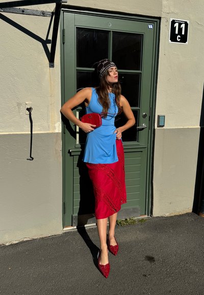 Top sin mangas de color azul claro, falda roja con intrincados detalles de encaje, zapatos de charol rojos y un pequeño bolso rojo; de pie junto a una puerta verde.