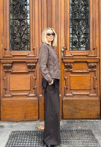 Pull en maille torsadée marron, pantalon large noir, chaussures noires ; debout à côté d'une porte en bois ornée avec des accents décoratifs en verre et métal.