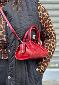 Κόκκινη τσάντα χρυσής κροκοδείλας με δύο κοντές λαβές και αποσπώμενο ώμο, resting σε ένα μαύρο ελαφρύ γιλέκο.