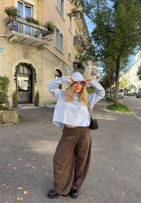 Młoda kobieta o kręconych włosach, ubrana w niebieską bluzę z kapturem i szerokie brązowe spodnie, stoi na słonecznej miejskiej ulicy z balkonami i drzewami.