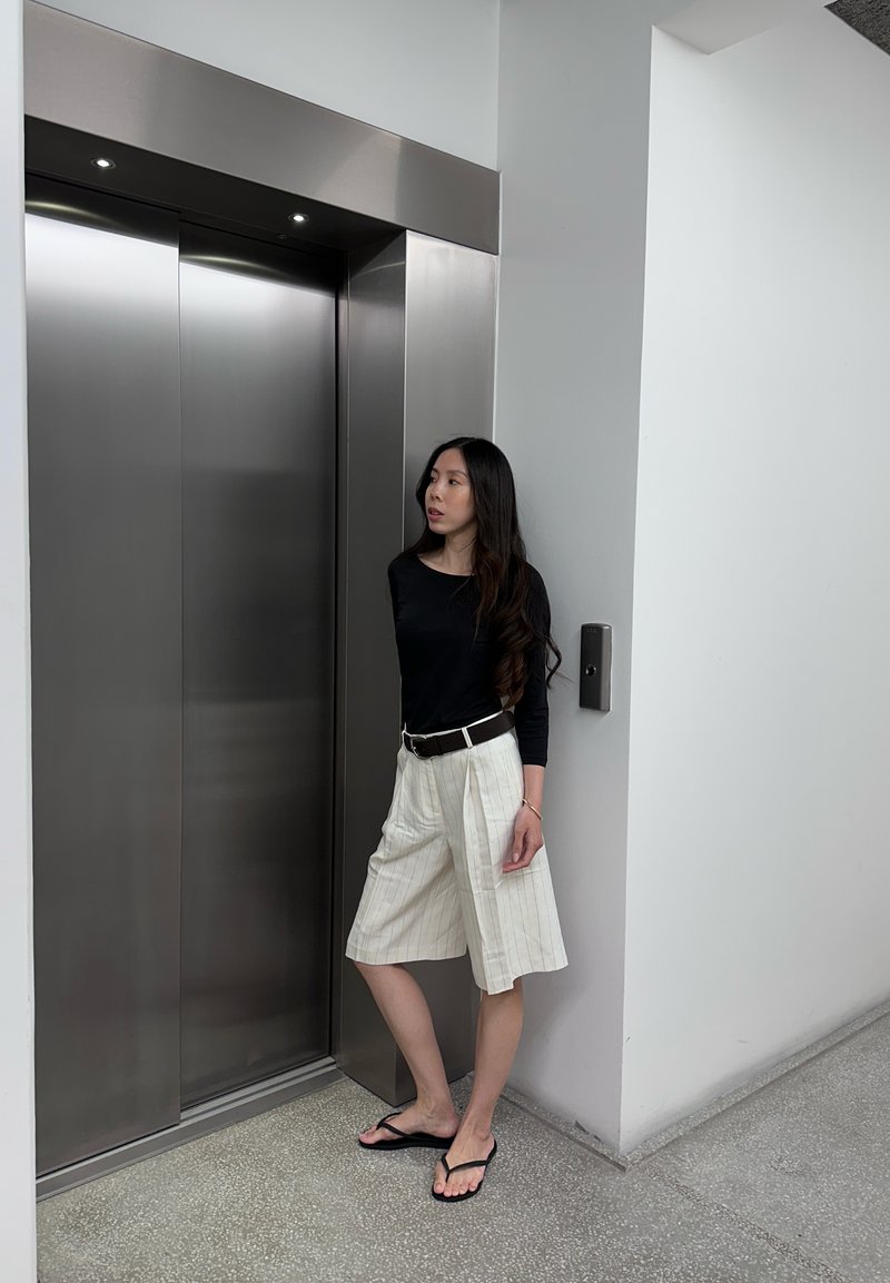Donna con top nero e pantaloncini bianchi in piedi vicino a porte di ascensore chiuse in acciaio inox in un ambiente interno minimalista.