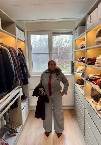 Mujer con suéter gris, corbata granate y pantalones anchos, de pie en un vestidor bien organizado con ropa doblada y abrigos colgados.