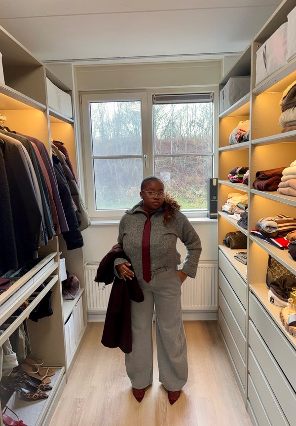 Frau im grauen Pullover, mit bordeauxroter Krawatte und weiten Hosen, steht in einem gut organisierten begehbaren Kleiderschrank mit gefalteter Kleidung und hängenden Mänteln.