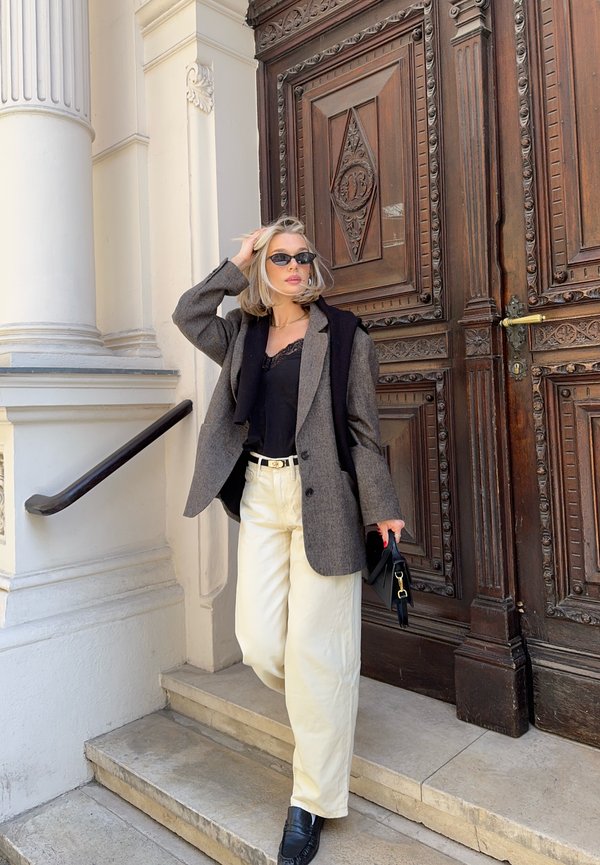 Frau mit Sonnenbrille, die einen grauen Blazer, ein schwarzes Top und weiße Hosen trägt, steht auf steinernen Stufen neben einer geschnitzten Holztür.