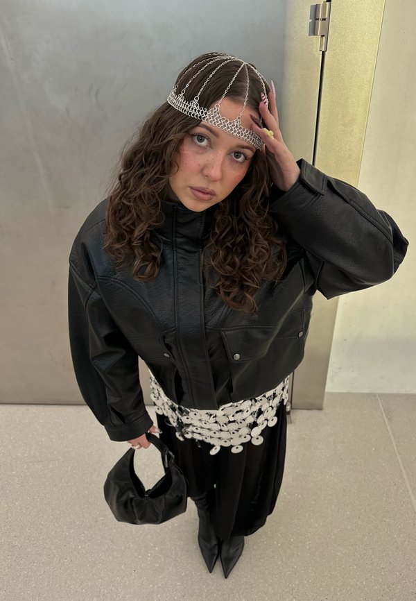 Młoda kobieta o kręconych włosach, nosząca łańcuszkową ozdobę na głowie, oversize'ową czarną skórzaną kurtkę, czarną spódnicę, biały pasek z dyskami, czarne botki, trzymająca czarną torebkę.