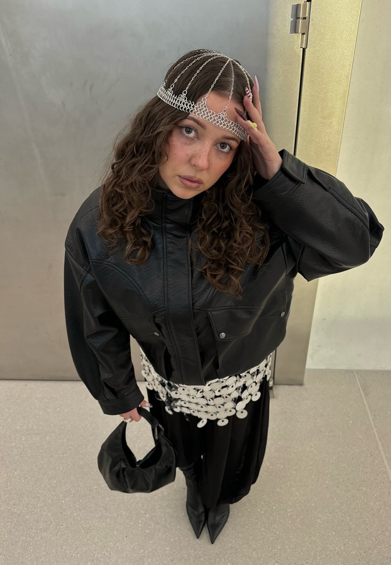 Junge Frau mit lockigem Haar, die einen Ketten-Haarschmuck trägt, übergroße schwarze Lederjacke, schwarzen Rock, weißen Gürtel mit Scheiben, schwarze Stiefel und eine schwarze Tasche hält.
