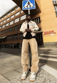 Homme en pantalon beige et blouson universitaire se tient sous un panneau de passage pour piétons sur un trottoir urbain près d'un restaurant et d'un bar.