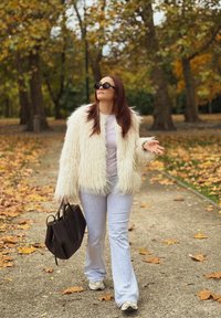 Krémová bunda z umelé kožušiny prehodená cez svetlú blúzku, sivé rozšírené nohavice a vzorované tenisky. Drží tmavú kabelku a je obklopená jesennými listami.