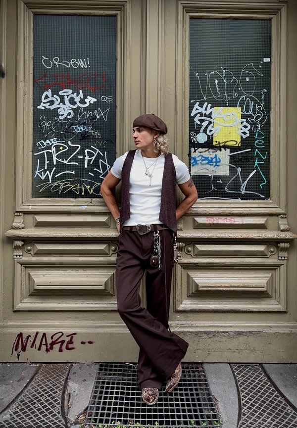 Pantaloni larghi marroni, t-shirt bianca, gilet marrone scuro e berretto abbinato. Gioielli d'argento notevoli e scarpe con fantasia contro uno sfondo di graffiti.