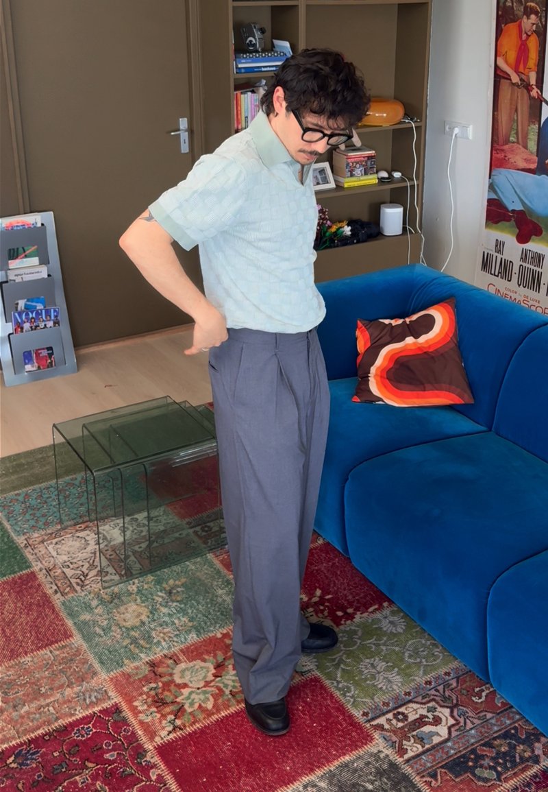 Homme avec lunettes et moustache ajustant son pantalon gris dans un salon coloré avec un canapé bleu et un tapis à motifs.