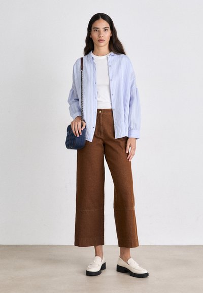 Camisa de botones a rayas azul claro sobre una camiseta blanca, combinada con pantalones de pierna ancha marrones y mocasines blancos; sostiene un bolso azul con estampado.