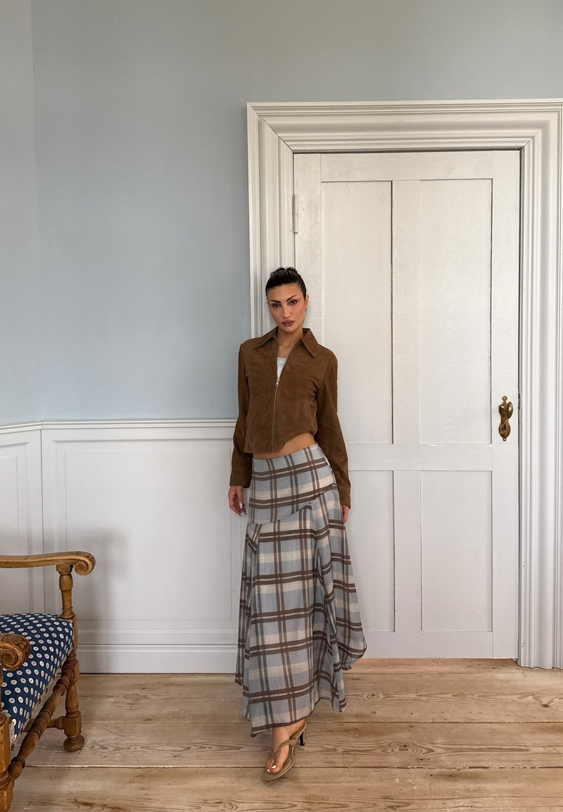 Femme debout à l'intérieur près d'une porte blanche, portant une veste marron zippée, une longue jupe à carreaux et des sandales, avec un fauteuil en bois à côté d'elle.