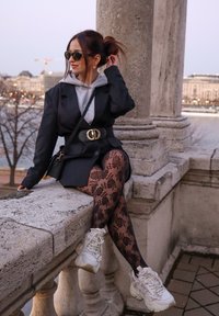 Blazer preto sobre um fato cinza, com meias-calças pretas de padrão floral e sapatilhas brancas chunky, contra um fundo de parapeito de pedra.