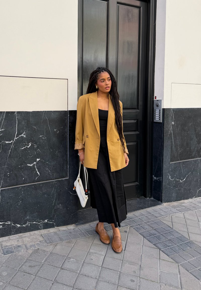 Femme aux longs cheveux tressés portant un blazer moutarde, une robe noire et des chaussures marron, debout contre un mur en marbre noir près d'une porte noire, tenant un sac blanc.