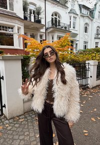 Puha fehér műszőrme kabát, barna crop top és bő barna nadrág. Kiegészítők között kerek napszemüveg. A háttér őszi lombkoronát ábrázol.