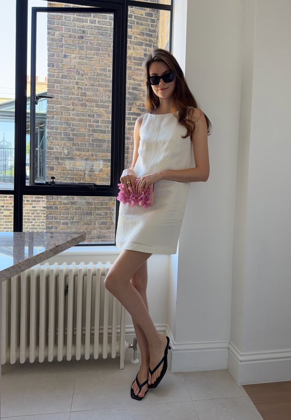 Femme en robe blanche sans manches avec des sandales noires se tient près d'une fenêtre en tenant une pochette rose texturée, portant des lunettes de soleil à l'intérieur.