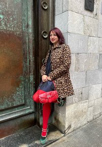 Leopardenmuster-Mantel, schwarzes Kleid, rote Strumpfhosen, Handtasche in Krokodillederoptik in Rot, grüne Plateauschuhe mit weißen Akzenten; steht neben einer strukturierten Wand.