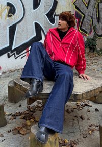 Červená bunda na zip s bílými pruhy, černý rolák, modré široké džíny a černé kotníkové boty, sedící na dřevěné lavici s graffiti na pozadí.