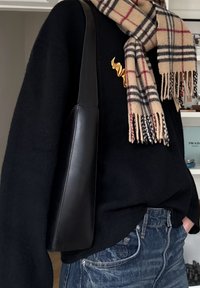 Personne portant un pull noir, un jean bleu, une écharpe à carreaux beige, et portant un sac à main en cuir noir à l'épaule à l'intérieur, avec une étagère en arrière-plan.