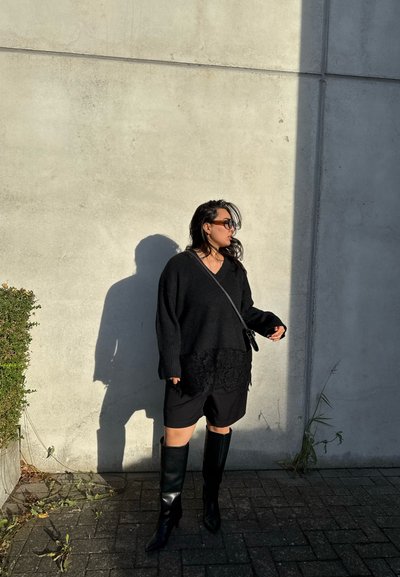 Mujer con suéter negro de gran tamaño, pantalones cortos negros, botas negras hasta la rodilla y gafas de sol, de pie junto a una pared de cemento con una sombra proyectada.