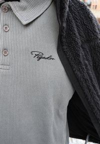 Hallikashallane soonikuga polo särk musta "Pegador" tikandiga, pruunide nuppudega ja osaliselt nähtav must fliisjope selle peal.