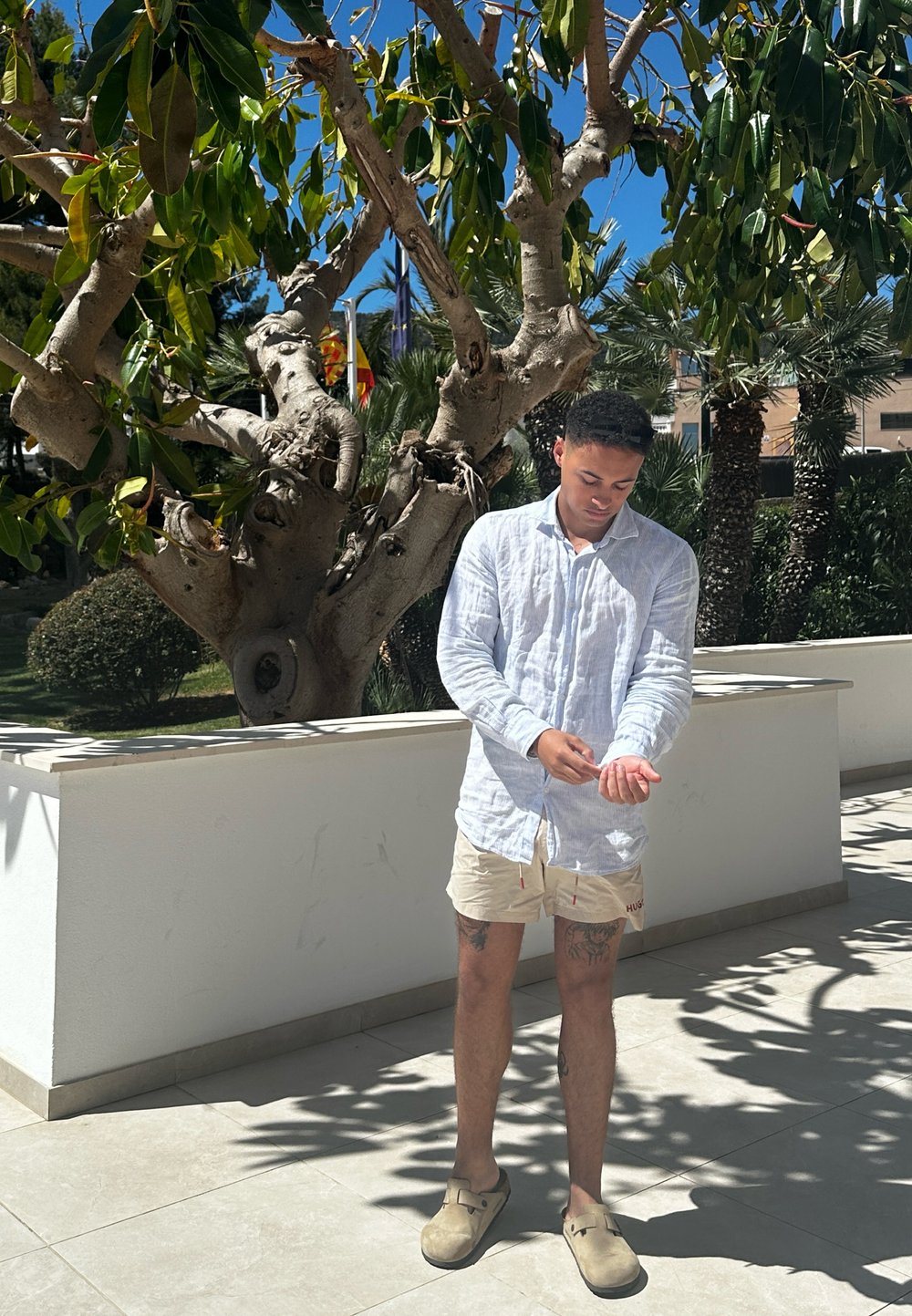 Camisa de linho azul claro, calções bege com cordões, e tamancos castanhos. De pé junto a uma parede branca com folhagem ao fundo.