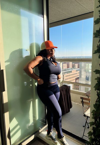 Top atlético azul marino y leggings, gorra naranja y blanca, de pie junto a una puerta de vidrio con vista a la ciudad, la luz del sol proyectando sombras, decoración moderna en el balcón.