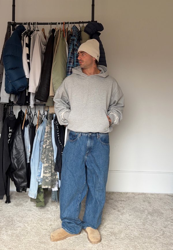 Homme portant un bonnet beige, un sweat à capuche gris, un jean bleu ample et des chaussons marron, debout les mains dans les poches à côté d'un portant à vêtements avec des vestes et des chemises.