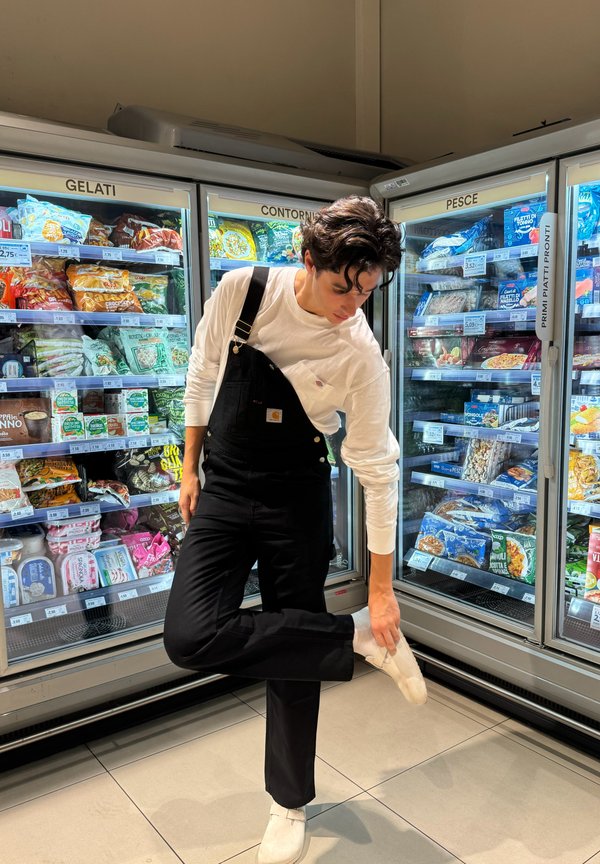 Pantalones de mono negros con una camiseta de manga larga blanca, de pie sobre una pierna. Sección de congeladores iluminada con alimentos envasados al fondo.