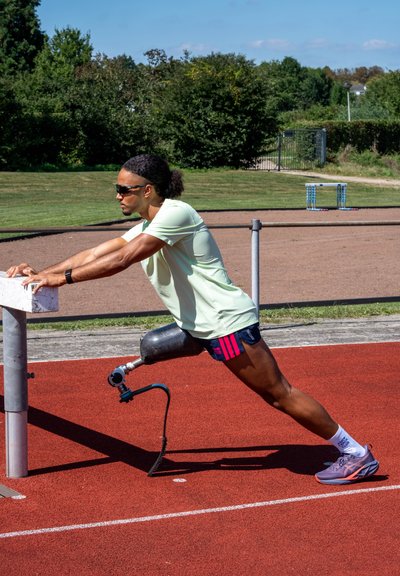 Atleta estirándose en una pista. Lleva una camiseta verde clara y pantalones cortos oscuros con acentos rosas. Usa una pierna prostética negra con herrajes azules.