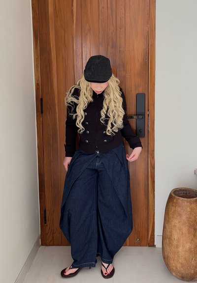 Persona con cabello rubio largo y rizado que lleva una gorra negra, una chaqueta negra abotonada, pantalones anchos azul oscuro y chanclas negras, de pie junto a una puerta de madera.